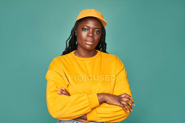 Femme afro-américaine dodue sans émotion en pull jaune et bonnet avec les bras croisés regardant la caméra tout en se tenant contre le mur bleu — Photo de stock