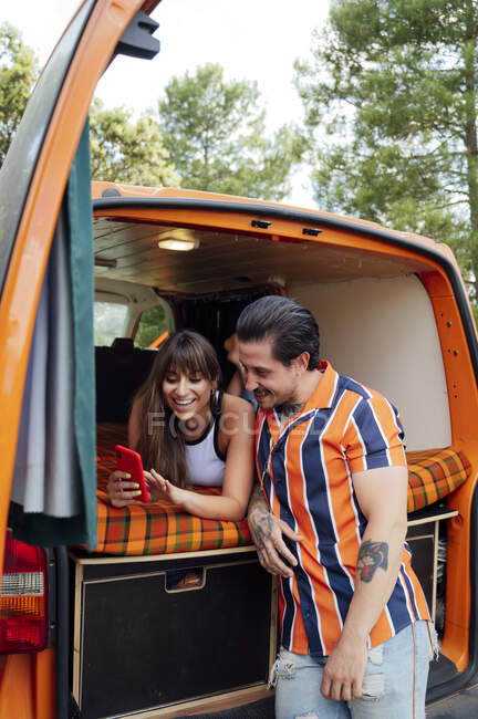Encantada pareja de viajeros sentados en furgoneta y tomando fotos en el teléfono inteligente mientras abrazan y disfrutan de la aventura de verano - foto de stock