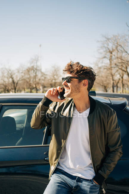 Щасливий чоловік в сонцезахисних окулярах говорить на мобільному телефоні, стоячи біля автомобіля в сонячний день — стокове фото