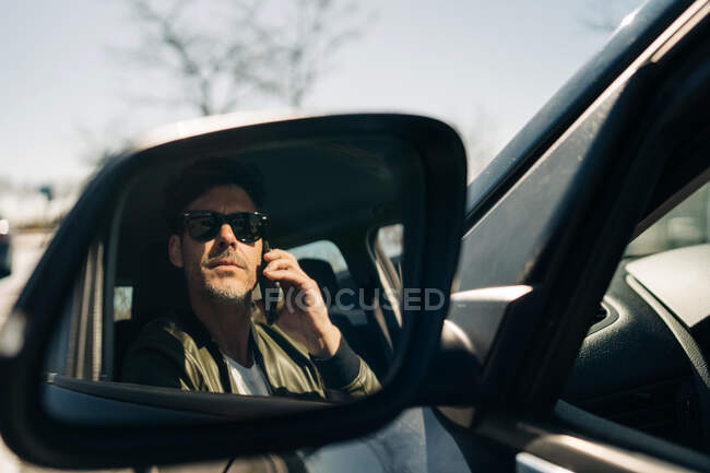 Homem barbudo em óculos de sol falando no celular enquanto reflete no espelho lateral do automóvel na luz solar — Fotografia de Stock
