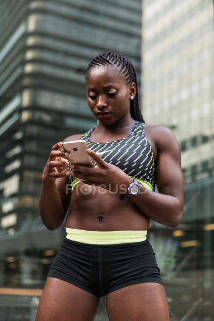 Afroamerikanerin in Sportkleidung surft auf modernem Smartphone, während sie beim Training im Freien auf verschwommenem Hintergrund der Stadtstraße steht — Stockfoto