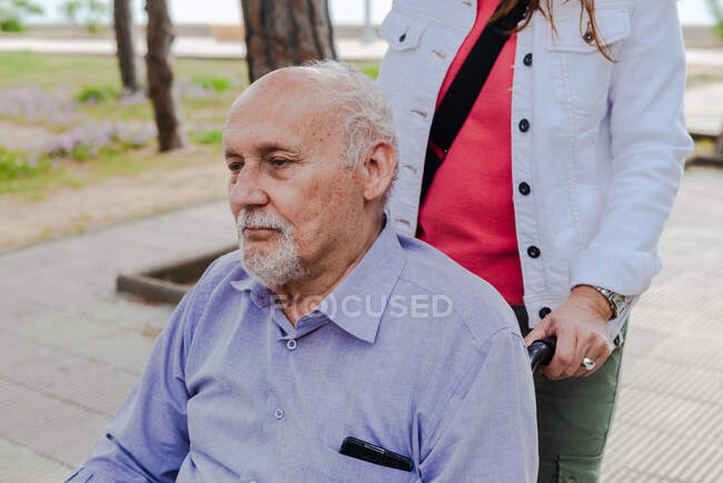 Врожай доросла дочка штовхає інвалідне крісло зі старим батьком під час прогулянки в парку влітку — стокове фото