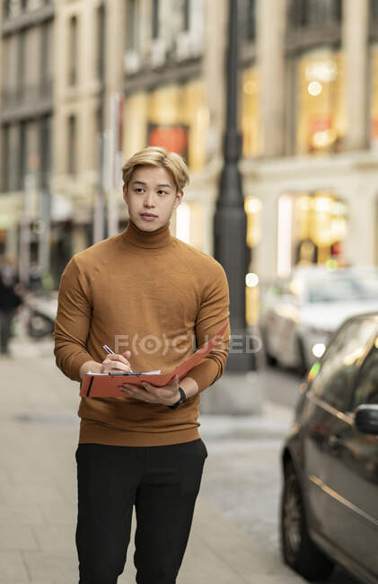 Focused ethnic male entrepreneur standing in street and writing on paper n folder — Stock Photo