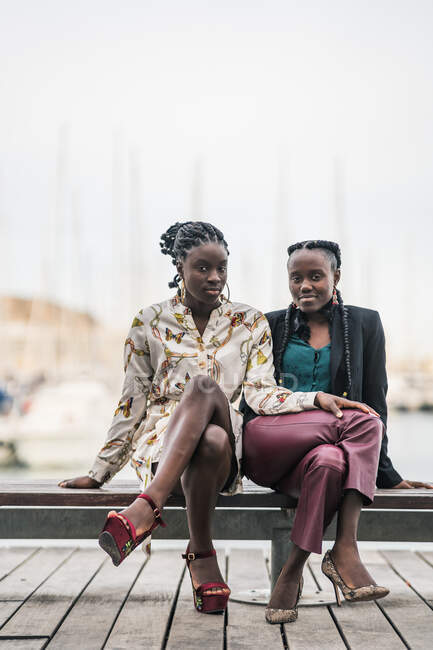Modische Afroamerikanerinnen sitzen bei hellem Tag auf einer niedrigen Holzbank im Park und schauen in die Kamera — Stockfoto