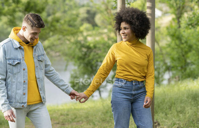 Encantado joven multiétnico y mujer tomados de la mano caminando por la costa del río en la naturaleza - foto de stock
