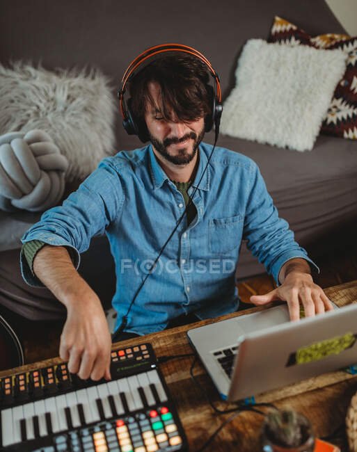 Joven feliz en auriculares usando sintetizador y portátil en la mesa en casa - foto de stock