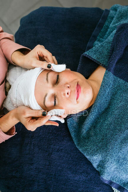 Top view of crop unrecognizable cosmetician using cotton pads and applying tonic on face of female customer lying on table in beauty salon during skincare procedure — Stock Photo