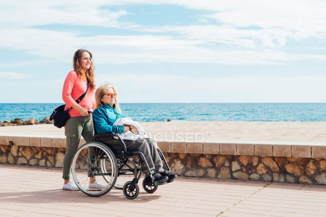 Вид сбоку на взрослую дочь, прогуливающуюся с пожилой матерью в инвалидной коляске по набережной у моря летом — стоковое фото