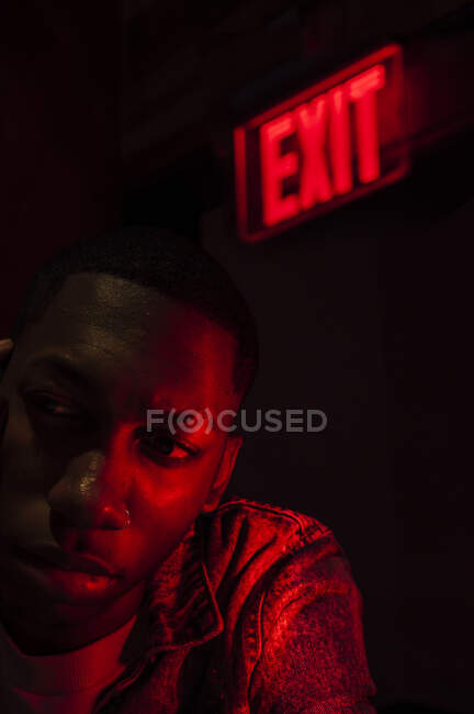 Афроамериканець, який дивиться у бік біля ілюмінованої таблетки Вихід над головою у червоному темному світлі. — стокове фото