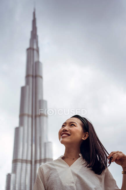 Low angle of smiling young Asian female in casual outfit looking away while  standing against modern high Burj Khalifa tower in Dubai in cloudy day —  construction, sight - Stock Photo | #517115458