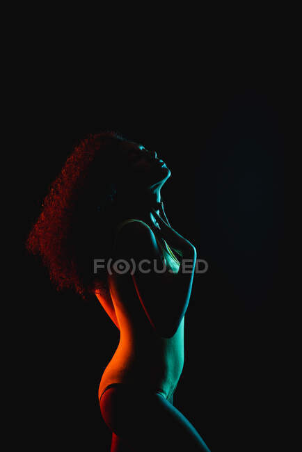 Vista lateral de una joven afroamericana con pelo rizado en ropa interior con la mano en la cintura tocando la barbilla con los ojos cerrados en el estudio de fondo negro - foto de stock