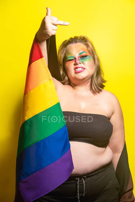 Rude plus size female with rainbow flag showing fuck you gesture while supporting LGBT against yellow background — Foto stock