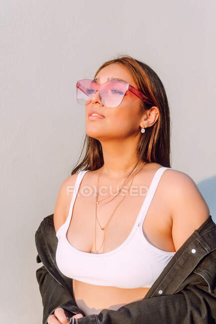 Уверенная молодая женщина в модной повседневной одежде и солнцезащитных очках, стоящих возле белой стены под солнцем — стоковое фото