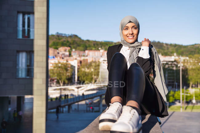 Mulher muçulmana encantada em desgaste elegante e lenço de cabeça sentado na rua e olhando para longe — Fotografia de Stock