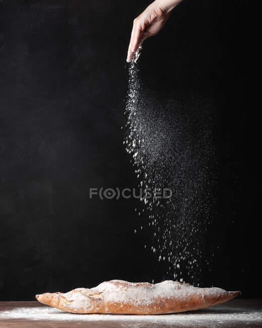 Erntehelfer anonymer Bäcker streut Weißmehl über frisch gebackenes Baguette-Brot vor schwarzem Hintergrund — Stockfoto