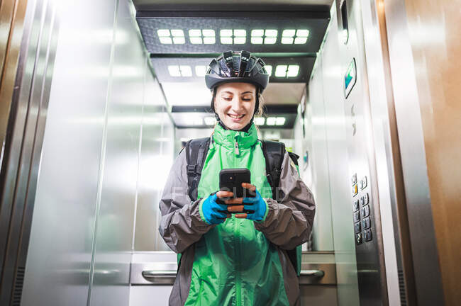 Baixo ângulo de correio feminino feliz em uniforme e capacete sorrindo e navegando celular no elevador ao fazer a entrega — Fotografia de Stock