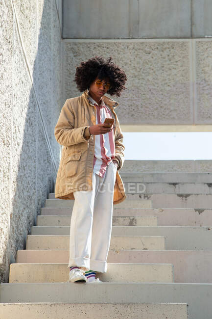 Basso angolo di etnia maschile in cappotto vintage con acconciatura afro messaggistica di testo sul cellulare su scala urbana — Foto stock