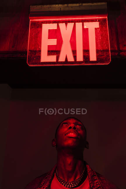 From below crop African American young male looking up on illuminated tablet Exit above head in red dark light — Stock Photo
