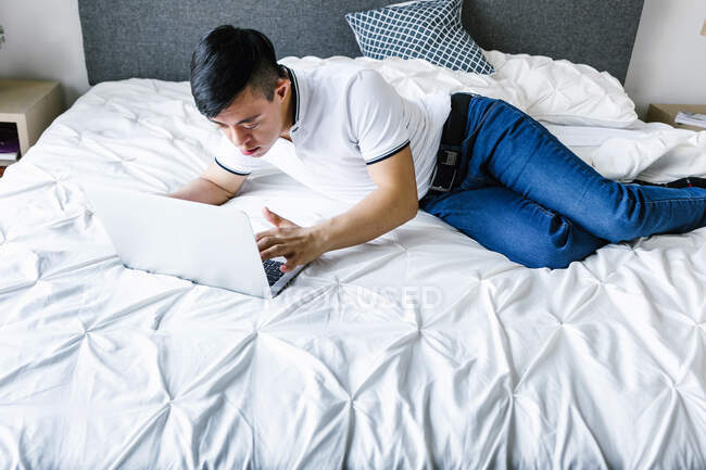 High angle of focused Latin teen boy with Down syndrome lying on bed and surfing Internet on netbook — Stock Photo