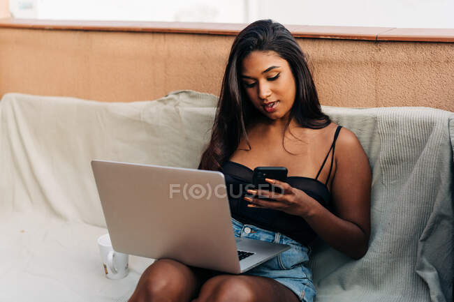 Молодая латиноамериканка сидит на диване рядом с ноутбуком, просматривает смартфон на балконе — стоковое фото