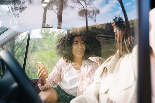 Through window of cheerful young Asian woman sitting on driver seat of camper vehicle while traveling with girlfriend through nature — Stock Photo