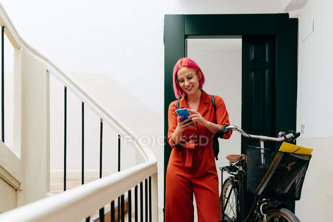 Молода стильна жінка в червоному костюмі з рюкзаком, використовуючи смартфон, стоячи з велосипедом на сходах — стокове фото