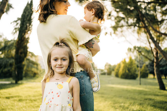 Мила дошкільнята посміхається і дивиться на камеру, обіймаючи матір з молодшою сестрою на руках протягом літнього дня разом у зеленому парку. — стокове фото