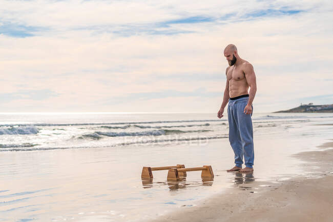 Vista lateral comprimento total do atleta masculino de pé se preparando para fazer o trabalho em bares paralelos na costa arenosa com ondas do oceano no fundo — Fotografia de Stock