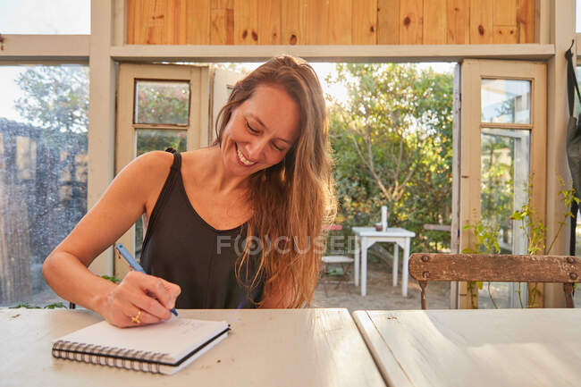 Niedriger Winkel der fröhlichen Gärtnerin sitzt am Tisch und schreibt in Notizblock im Gewächshaus — Stockfoto