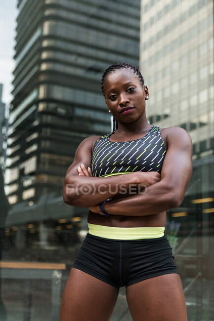 Позитивна чорна жінка в спортивному одязі тримає руки схрещеними і дивиться на камеру, стоячи на розмитому тлі міської вулиці — стокове фото