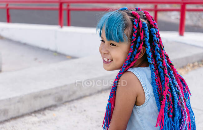 Criança étnica bonita alegre com tranças coloridas sentadas na escada de concreto enquanto olha para longe à luz do dia — Fotografia de Stock