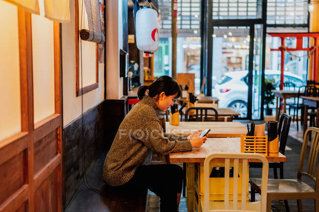 Asian lady in casual sweater using mobile phone at counter in traditional ramen bar — Stock Photo