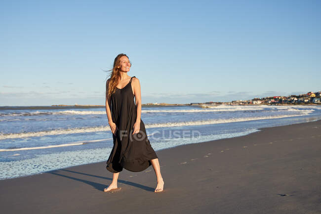 Cuerpo completo sonriente hembra en vestido de verano de pie en la orilla del mar de arena y mirando hacia otro lado - foto de stock