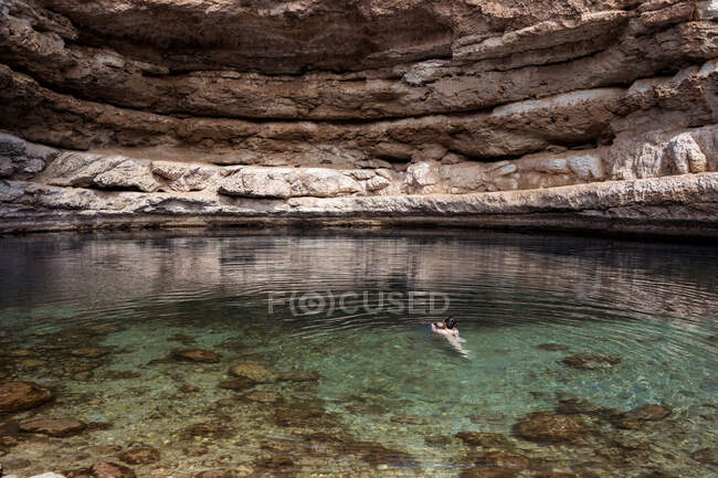 Mujer anónima relajada flotando en el agua transparente del sumidero de Bimmah rodeada de rocas ásperas durante el viaje en Omán - foto de stock