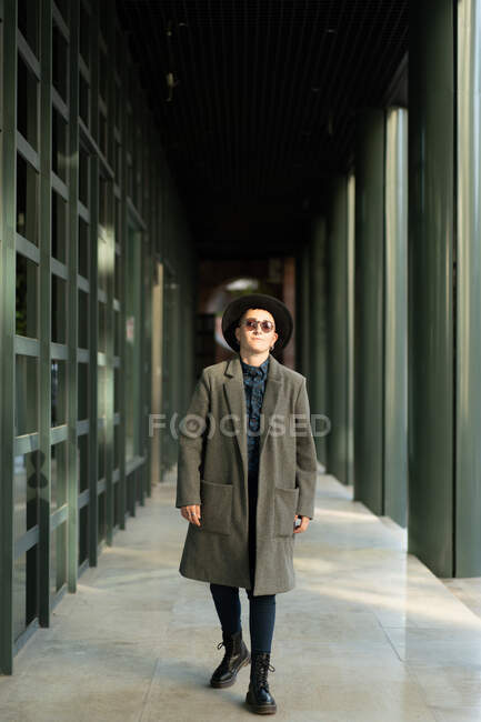 Transgênero pessoa de casaco e óculos de sol andando no piso de azulejos do edifício no dia ensolarado — Fotografia de Stock