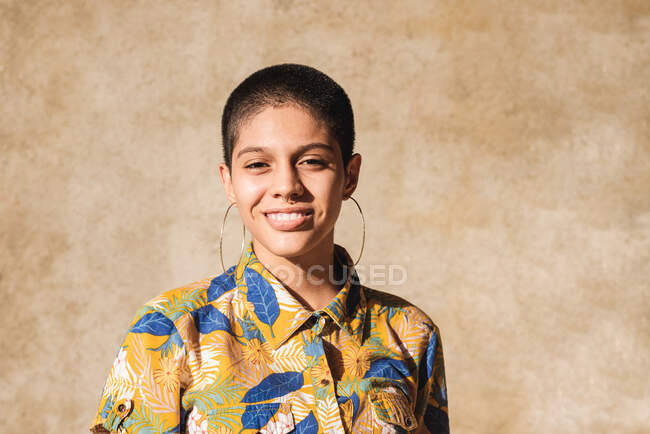 Contenu jeune femme bisexuelle ethnique en boucles d'oreilles regardant la caméra sur fond beige à l'extérieur — Photo de stock