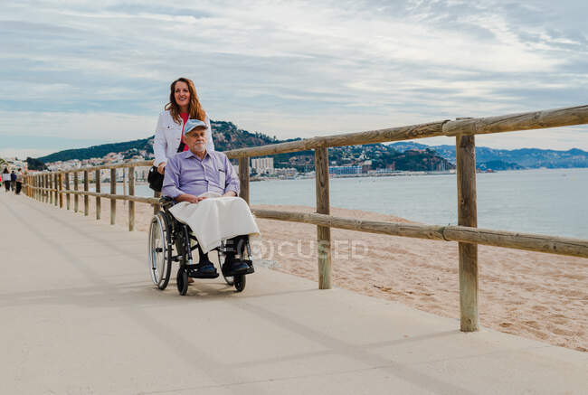 Delighted adult daughter pushing wheelchair with senior father and enjoying stroll along promenade near sea — Stock Photo
