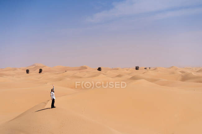 Smiling young woman in casual clothes standing on sandy dune against desert during travel in Emirates and waving hand — Stock Photo