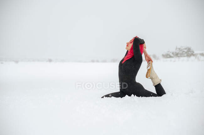 Спокойная женщина с розовыми волосами сидит в Eka Pada Raja Kapotasana на снежном зимнем поле и практикует йогу — стоковое фото