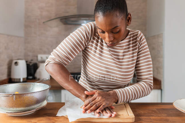 Joven mujer afroamericana aplastando plátano fresco en la tabla de cortar mientras prepara patacones en casa - foto de stock