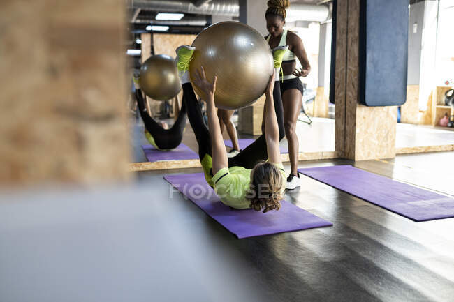Unrecognizable sportsman with Pilates ball exercising on mat against black female athlete and mirror in gymnasium — Stock Photo