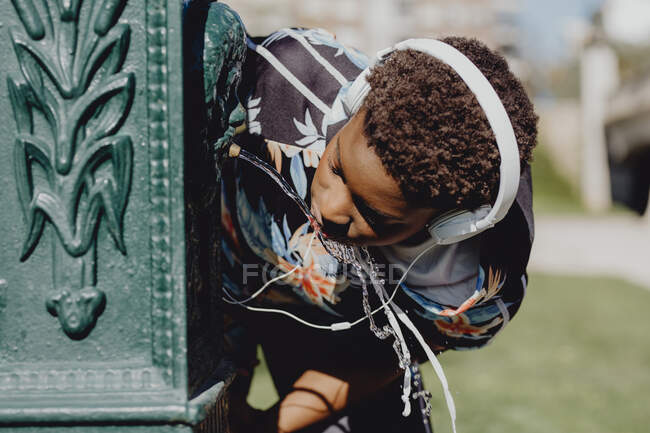 Joven fitness Afroamericana bebiendo agua de una fuente mientras escucha música en un día soleado en la calle de la ciudad - foto de stock