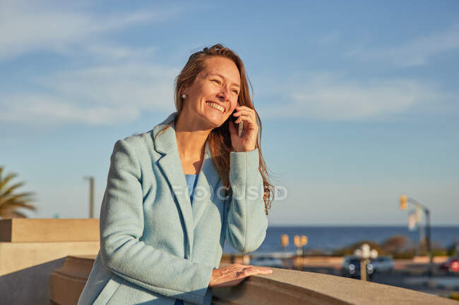 Улыбающаяся взрослая женщина в теплом пальто разговаривает по телефону, опираясь на забор возле океана на городской улице в солнечный день — стоковое фото