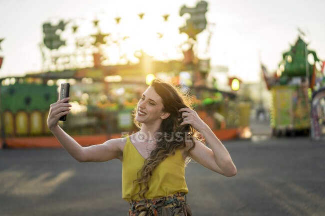 Conteúdo fêmea fazendo auto-retrato no celular enquanto se diverte no parque de diversões à noite no verão — Fotografia de Stock