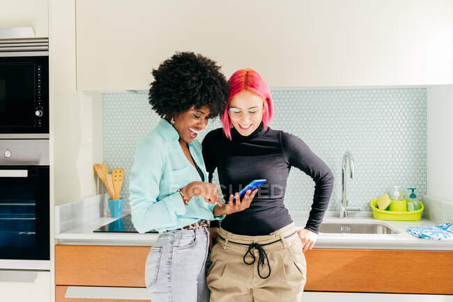 Веселі молоді багаторасові друзі-жінки в модному вбранні, використовуючи мобільний телефон та обговорюючи новини в соціальних мережах, стоячи на домашній кухні — стокове фото