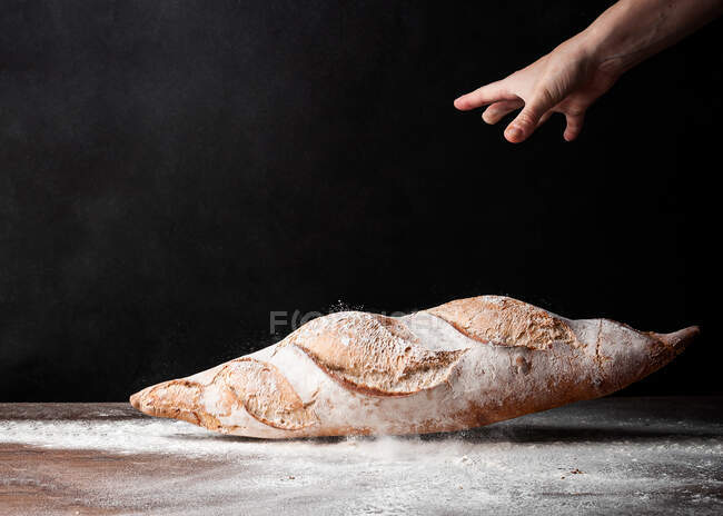 Crop anonymous baker dropping baked bread dusted with flour on the table against black background — Stock Photo