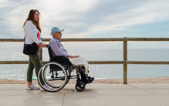 Восхитительная взрослая дочь толкает инвалидное кресло со старшим отцом и наслаждается прогулкой вдоль набережной возле моря — стоковое фото