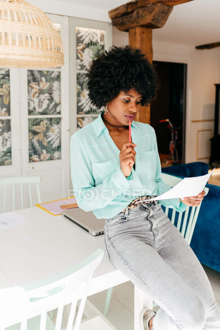 Moderna donna afro-americana freelance di successo in abito elegante con capelli afro seduta a tavola e documento di lettura a casa — Foto stock