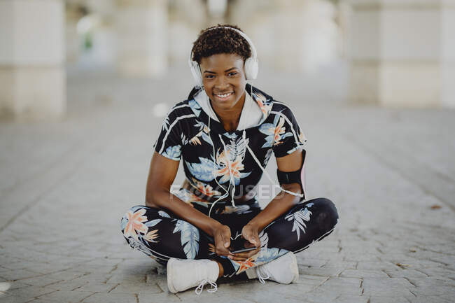 Young fitness African American woman resting and stretching legs while listening to music after running on city street — Stock Photo