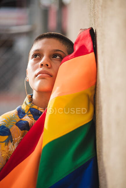Joven mujer étnica soñadora con bandera colorida y pelo corto mirando hacia arriba contra la pared sobre fondo borroso - foto de stock